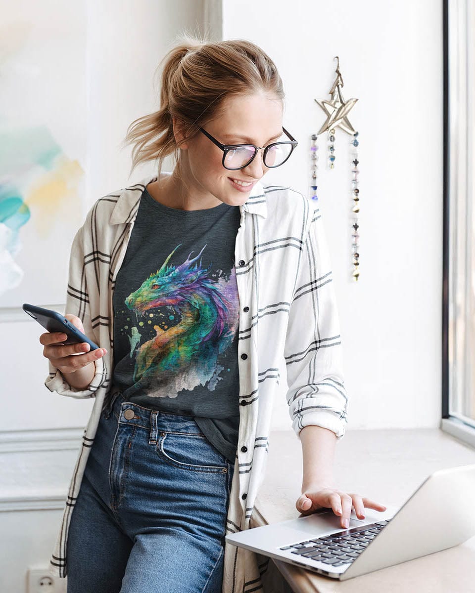 Creative woman wearing Chromatic Sentinel shirt featuring watercolor art, holding her phone and working on a laptop.