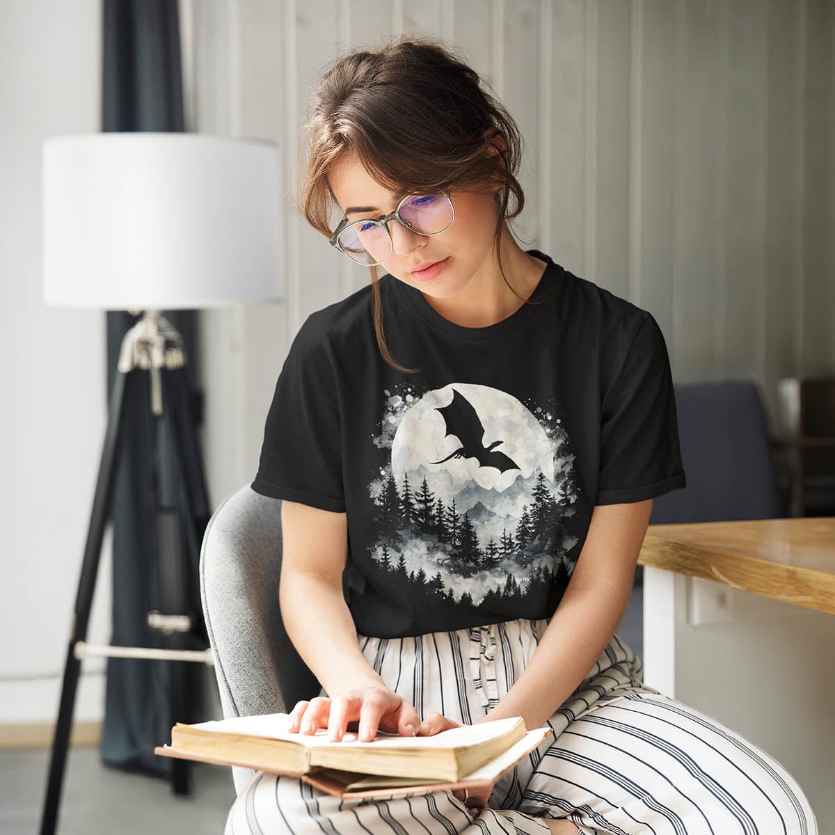 Woman reading a book while wearing a Great Dragon Shirt featuring a dragon silhouette flying past a full moon and mountains.