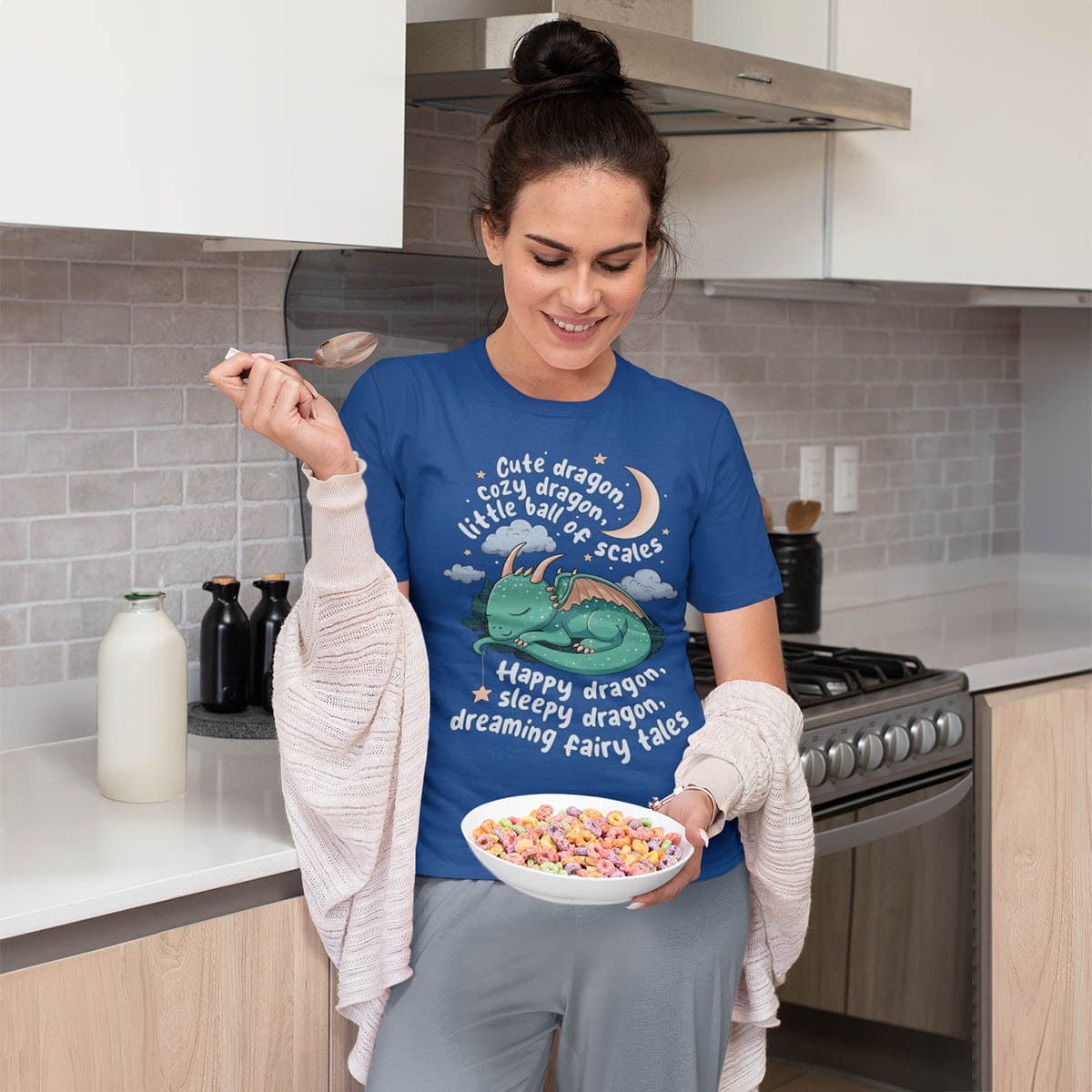 Woman lounging in a kitchen wearing a Happy Dragon Shirt featuring a cute sleeping dragon and poem, eating a bowl of cereal.