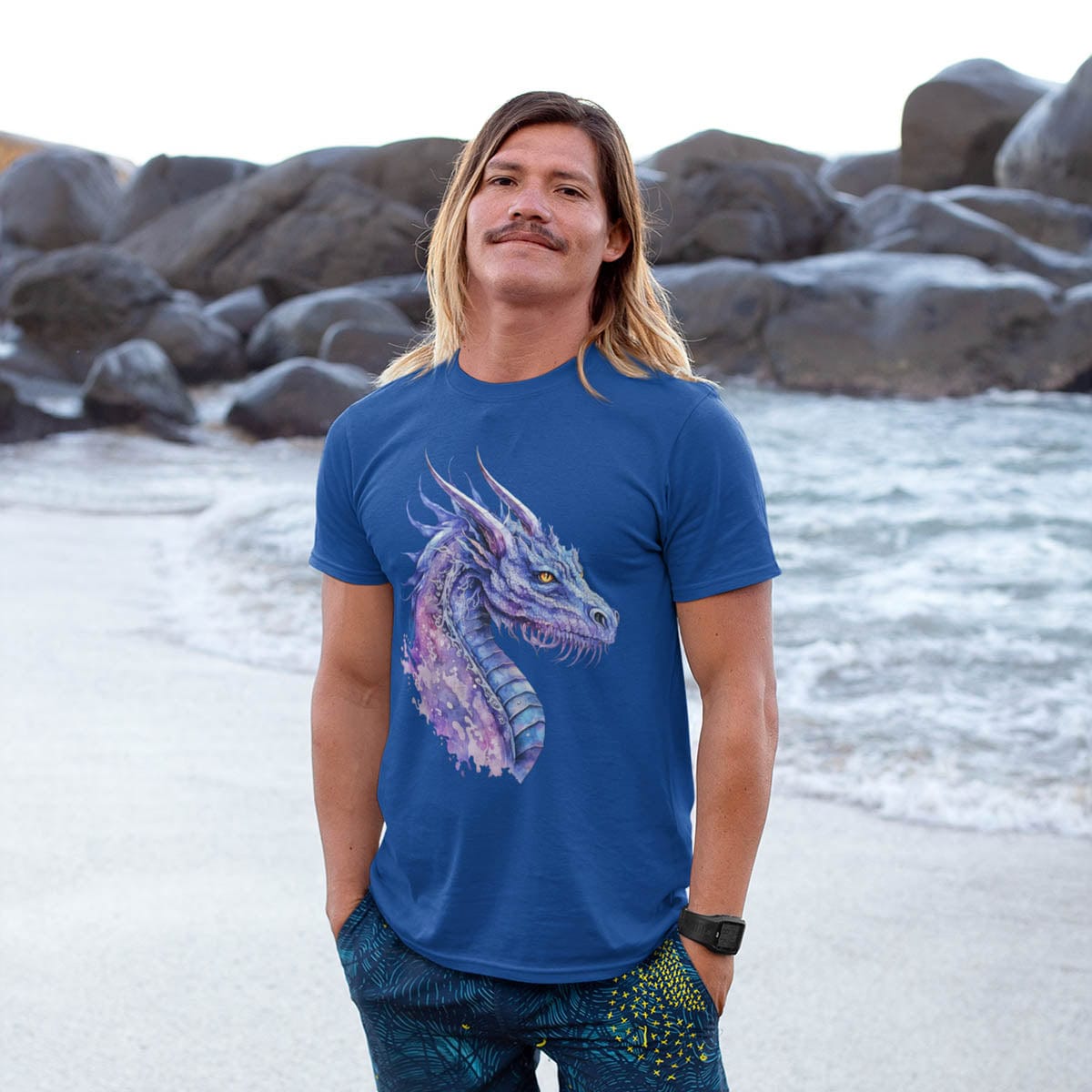 Man standing on a rocky beach wearing a Blue and Purple Dragon Shirt featuring ethereal watercolor art of an aquatic dragon.